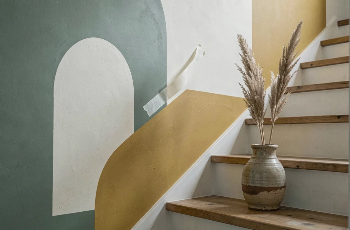 Détail d'une peinture murale graphique color-blocking vert sauge et moutarde sur mur d'escalier avec pot en céramique vintage
